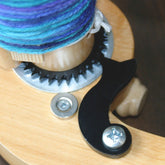 A close-up view of a Kromski Harp Forte spinning wheel mechanism reveals a section of the wheel with a gear and yarn in shades of blue and purple. A black hook and metal screw are visible on the wooden surface, highlighting the fine craftsmanship enhanced by the Kromski Harp Forte Ratchet/Pawl Upgrade Kit from Kromski North America.