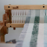 A wooden Kromski Weaver's Choice - Rigid Heddle Reed from Kromski North America, with a partially woven fabric featuring light blue and white stripes. The white threads are aligned and appear taut, interspersed with some blue threads. The background is out of focus, showcasing this loom as an excellent outlet for weaving creativity with its adjustable dent heddle.