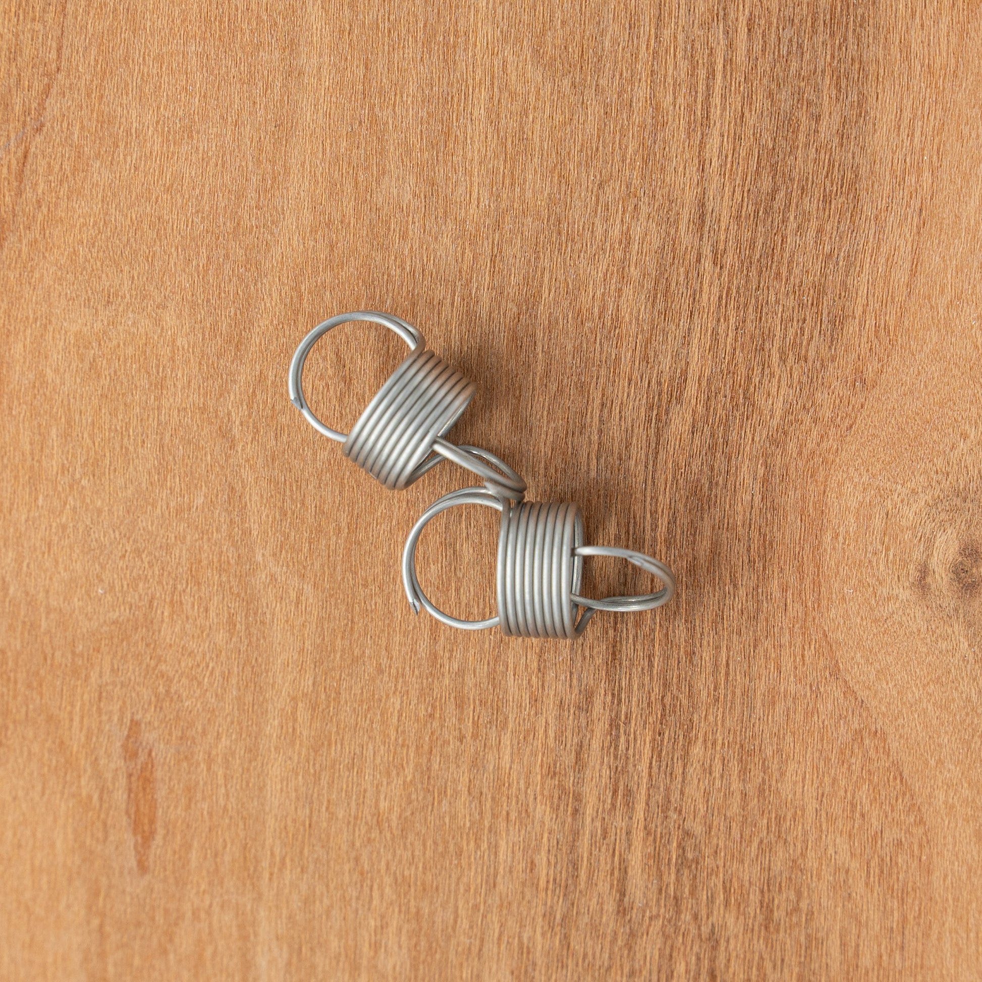 Two Kromski Spinning Wheel Springs, featuring coiled silver wires with loops, are placed on a wooden surface, echoing the intricate designs of Kromski North America's spinning wheels.