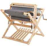 A Schacht Wolf Loom from Schacht Spindle Co., showcasing a partially woven gray fabric, features a hand crank on the right side along with numerous parts to adjust the warp and weft threads. The loom's sturdy frame includes a stand equipped with a wooden foot pedal, add-on reed slots, and versatility-enhancing Schacht Wolf Loom 4-Treadle Add-On Kits—ensuring flexibility for your weaving projects.