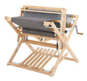 A Schacht Spindle Co. Schacht Wolf Loom 4-Treadle Add-On Kit, featuring an upright wooden frame with a large gray textile partially woven. The loom, made from light-colored sturdy wood, includes side supports and various mechanical parts such as a hand crank for added weaving versatility.