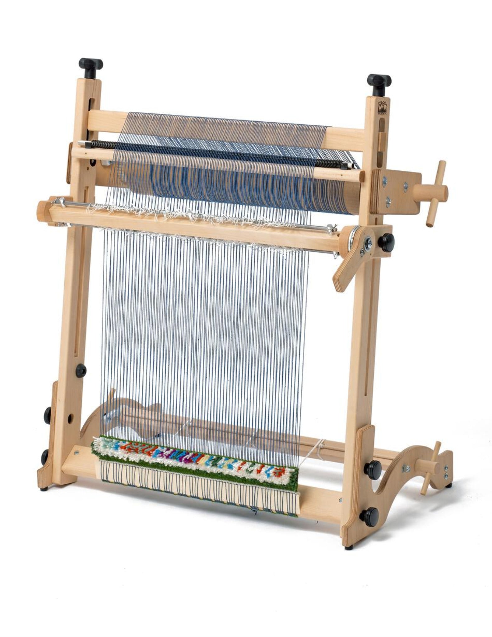 An Arras 20" Tapestry Loom Beam Kit from Schacht Spindle Co., set up with threads ready for weaving. The loom features various knobs and mechanisms for adjusting tension, employing a traditional warping technique. A section of colorful woven fabric is visible at the bottom. The plain white background highlights the loom's structure.