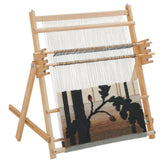 A Schacht Spindle Co. Schacht Tapestry Loom, 25" holding a tapestry in progress. The tapestry features a partially completed design with dark silhouettes of branches and leaves set against a lighter background. The loom, equipped with a tension beam, stands on a wooden frame with adjustable supports.