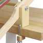 A Schacht 15" Table Loom Floor Stand from Schacht Spindle Co. is securely clamped to the edge of a wooden table using a metal C-clamp. The loom stand features multiple colored threads and a wooden frame held together with metal nuts and bolts, providing a sturdy installation for your weaving projects.