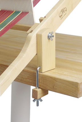 A Schacht 15" Table Loom Floor Stand from Schacht Spindle Co. is securely clamped to the edge of a wooden table using a metal C-clamp. The loom stand features multiple colored threads and a wooden frame held together with metal nuts and bolts, providing a sturdy installation for your weaving projects.