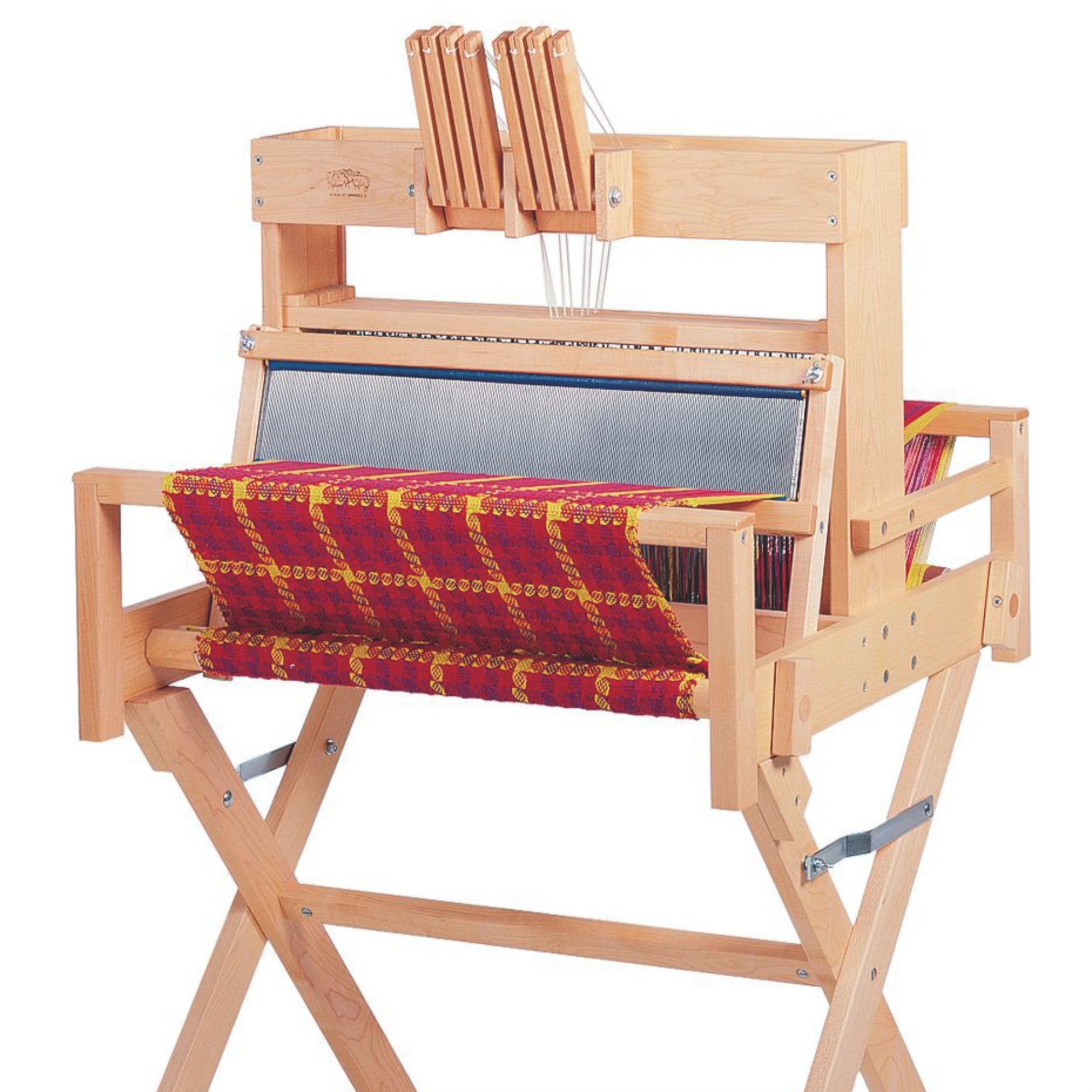 A Schacht Spindle Co. 15" Table Loom Floor Stand holds a wooden tabletop weaving loom, which features a half-finished fabric with red and yellow checkered patterns. The intricate design showcases various levers and heddles on the top and front of the loom.