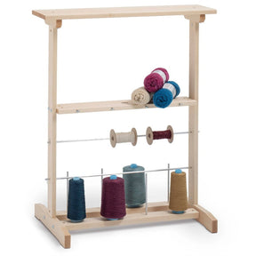 The Schacht Winding Station by Schacht Spindle Co. features two horizontal shelves that can accommodate numerous spools of colorful thread on the top shelf and three larger cones in maroon, blue, and beige on the lower shelf. A rod positioned between the shelves is perfect for holding smaller spools. This stand is ideal for organizing your winding tools, such as ball winders or swifts.