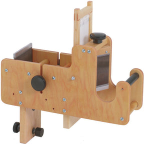 The Schacht Tension Box by Schacht Spindle Co. is a compact and functional wooden tabletop drum carder featuring metal components and rotating knobs. It includes a handle for manual operation, an adjustable tension knob for precision, metal carding cloth for processing fiber, and various screws for adjustment and stability.