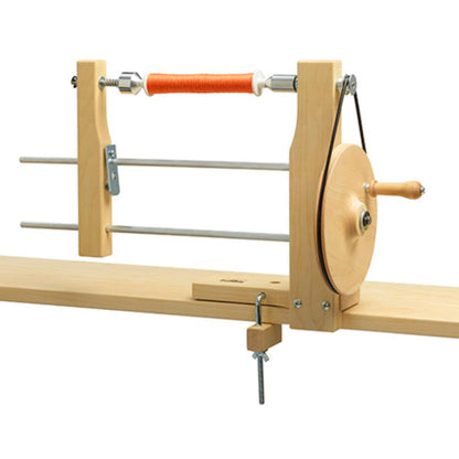 The Schacht Hand Bobbin Winder by Schacht Spindle Co. is a wooden device equipped with a hand crank and horizontal metal rods, designed for winding yarn into balls from skeins. It can be clamped to the edge of a table and features an orange yarn wound around its central spindle.