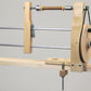 The Schacht Double Ended Hand Lace Bobbin Winder by Schacht Spindle Co. is a combination of wood and metal, mounted on the edge of a table. It includes two horizontal metal rods and a crank handle on the right side, with a small wooden clamp securing it firmly in place. The bobbin winder is set against a neutral backdrop with a plain background.