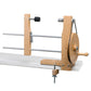 The Schacht Double Ended Hand Lace Bobbin Winder by Schacht Spindle Co. is clamped to the edge of a tabletop. The apparatus features a manual crank handle on the right, a circular wooden wheel, and metal rods for holding and winding yarn. A black tensioner is mounted on the vertical support.