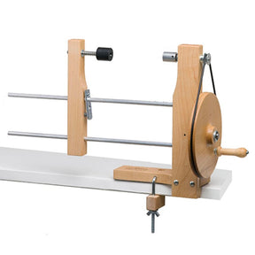 The Schacht Double Ended Hand Lace Bobbin Winder by Schacht Spindle Co. is clamped to the edge of a tabletop. The apparatus features a manual crank handle on the right, a circular wooden wheel, and metal rods for holding and winding yarn. A black tensioner is mounted on the vertical support.