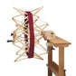 A Schacht Ultra Umbrella Swift yarn winder from Schacht Spindle Co., holding a skein of red yarn, is attached to a wooden table. The swift is fully extended, accommodating skeins up to 93 inches, and the table features a clamp for secure attachment. The overall setup, complete with an optional counter, is ready for winding yarn into balls.