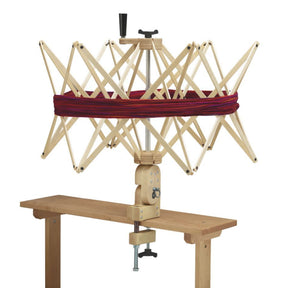 A Schacht Spindle Co. Schacht Ultra Umbrella Swift is clamped to a table, with red and purple yarn wrapped around its expandable arms. Designed to handle skeins up to 93 inches, the swift features a turn handle on top and additional supporting parts for secure positioning, with an optional counter available for precise measurements.