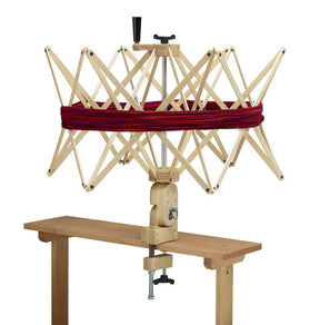 A Schacht Ultra Umbrella Swift by Schacht Spindle Co., designed for winding yarn into skeins up to 93", is clamped to a wooden table. The swift is extended with red yarn wrapped around it, ready for use. A black knob is visible on top for adjustment against a plain white background.