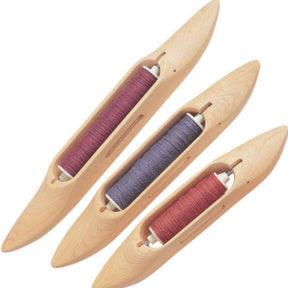 Three Schacht Boat Shuttles from Schacht Spindle Co. are arranged diagonally, each holding a spool of thread in different colors: one with red thread, one with purple thread, and one with orange thread. The shuttles are smooth and elongated, featuring a central slot for the thread.