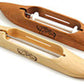 The image features two Schacht Boat Shuttles from Schacht Spindle Co., traditional wooden tools used in weaving. One shuttle is light-colored, while the other has a darker hue. Both are elongated in shape with pointed ends and include a central compartment designed to hold thread or yarn. Intricate decorative designs are etched on the surfaces of each shuttle.