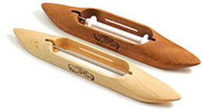 The image features two Schacht Boat Shuttles from Schacht Spindle Co., traditional wooden tools used in weaving. One shuttle is light-colored, while the other has a darker hue. Both are elongated in shape with pointed ends and include a central compartment designed to hold thread or yarn. Intricate decorative designs are etched on the surfaces of each shuttle.