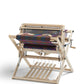 A Schacht 26" Baby Wolf Floor Loom from Schacht Spindle Co. displays a colorful woven fabric in progress, featuring various shades and patterns. The loom includes multiple levers and beams, with treadles at the bottom for operating the weaving mechanism. It is set against a plain white background.