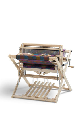 A Schacht 26" Baby Wolf Floor Loom from Schacht Spindle Co. displays a colorful woven fabric in progress, featuring various shades and patterns. The loom includes multiple levers and beams, with treadles at the bottom for operating the weaving mechanism. It is set against a plain white background.