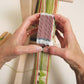 Hands are manipulating a set of Schacht Card/Tablet Weaving Cards (25/pack) from Schacht Spindle Co., arranged in a stack, which are used for card weaving. Green, white, and red threads are visible, forming a patterned strip of woven fabric. A wooden loom structure is partially visible, supporting this ancient craft of card weaving.