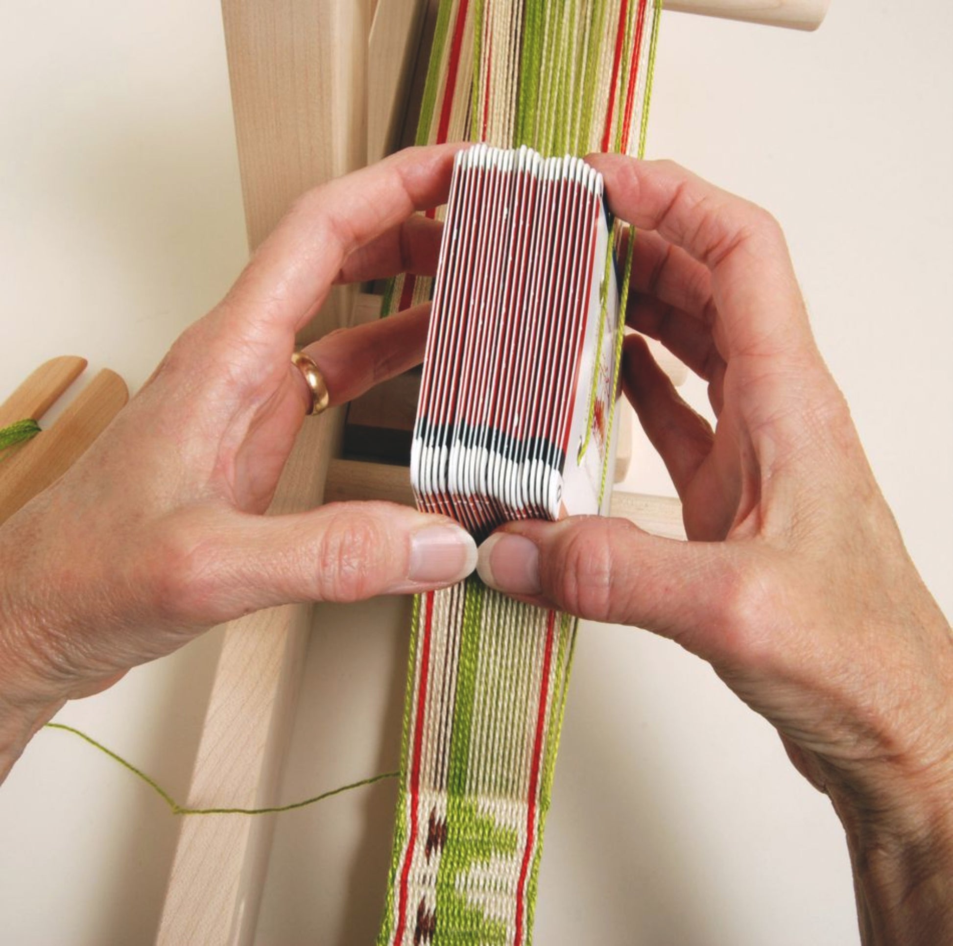 Hands are manipulating a set of Schacht Card/Tablet Weaving Cards (25/pack) from Schacht Spindle Co., arranged in a stack, which are used for card weaving. Green, white, and red threads are visible, forming a patterned strip of woven fabric. A wooden loom structure is partially visible, supporting this ancient craft of card weaving.