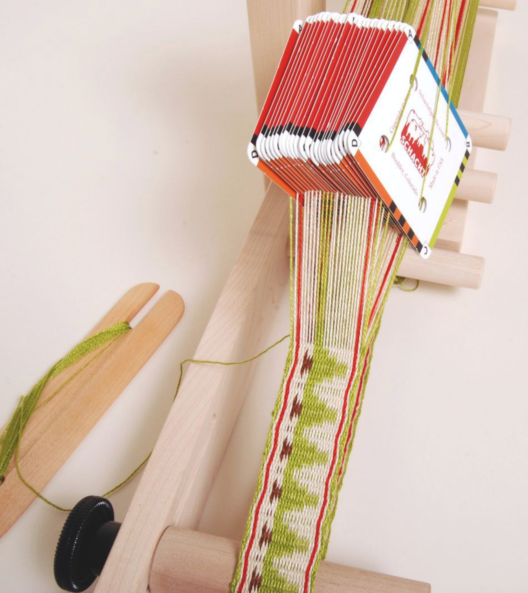 A weaving loom with a partially completed, intricate textile. The design features green, white, and red threads forming a patterned strip. A stack of Schacht Card/Tablet Weaving Cards (25/pack) by Schacht Spindle Co., each with holes for card weaving, hangs over the loom to aid in crafting historic patterns. A wooden shuttle is visible on the side.