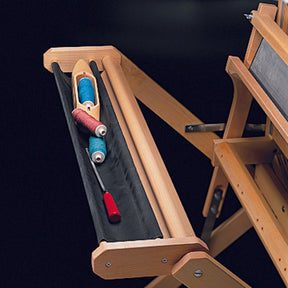 Close-up of a Schacht Spindle Co. Floor Loom Trap with a black tray. The tray holds a red-handled tool and three spools of thread in blue, white, and pink. The loom's wooden frame is visible, showcasing its structure and craftsmanship. The dark background highlights the loom's accessories.