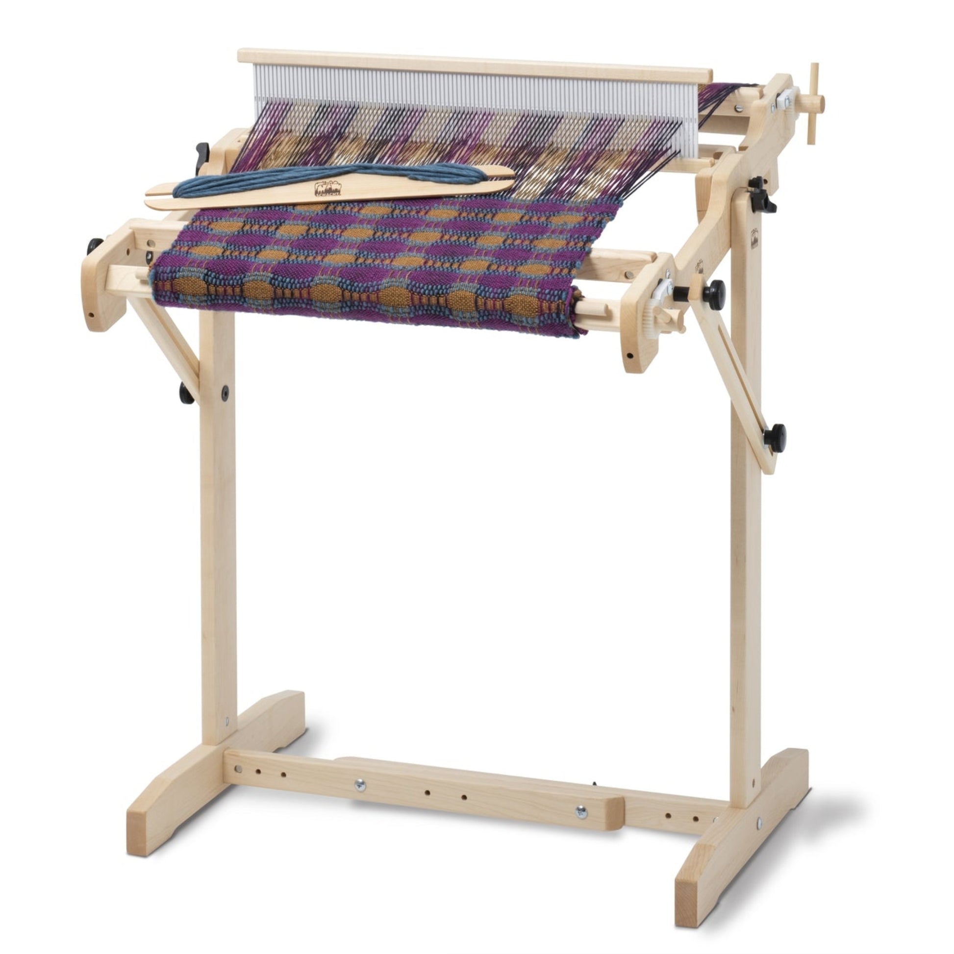 A Schacht Trestle Floor Stand for Flip Looms by Schacht Spindle Co. stands on a sturdy frame. The loom features a partially woven fabric with a vibrant purple and orange checkered pattern. Various components, including the shuttle, are visible and positioned within the adjustable width supports of the Flip and Tapestry Looms.