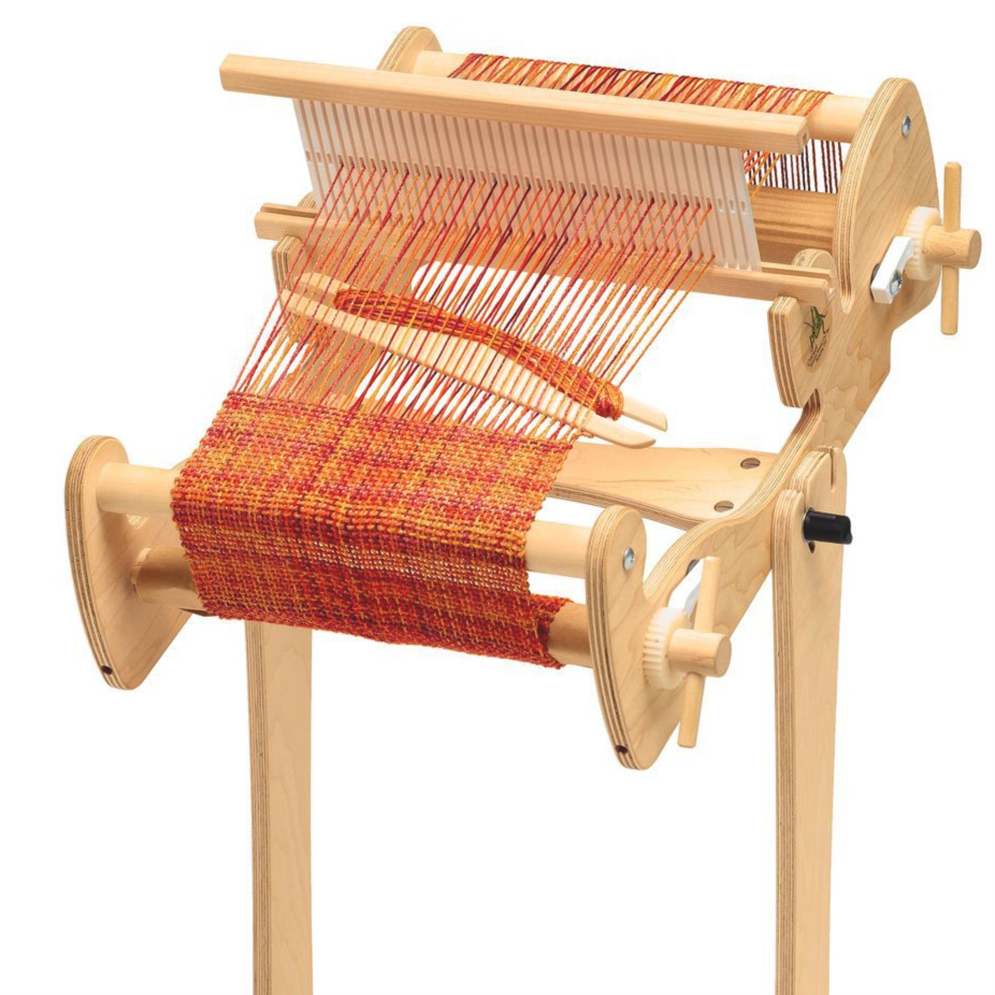 A high-quality apple ply tabletop weaving loom, called a Cricket Loom, is adorned with red and orange threads and features a simple design including a frame, roller, and heddle as it actively weaves cloth. The loom is securely placed on the Schacht Cricket Floor Stand by Schacht Spindle Co., made from light-colored wood.