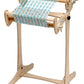 A high-quality Schacht Cricket Floor Stand from Schacht Spindle Co. stands upright, featuring a partially woven fabric in multicolored threads. The loom has a sturdy frame with crank handles and rollers, allowing for the creation of intricate woven patterns.