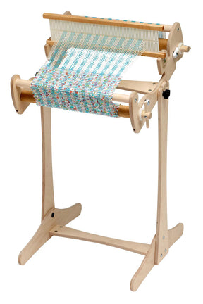 A high-quality Schacht Cricket Floor Stand from Schacht Spindle Co. stands upright, featuring a partially woven fabric in multicolored threads. The loom has a sturdy frame with crank handles and rollers, allowing for the creation of intricate woven patterns.