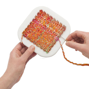 Two hands are shown using the Schacht Zoom Loom 4" x 4" from Schacht Spindle Co. to create a colorful, multi-hued orange textile. A needle guides the yarn through the pegs on the rounded square-shaped white loom, crafting an intricate pattern.