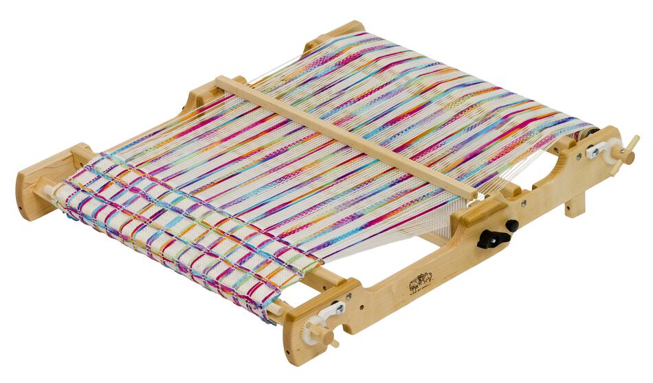 The Schacht Flip Folding Rigid Heddle Loom by Schacht Spindle Co. has colorful threads in shades of blue, red, yellow, and white stretched across it. The loom appears to be in use, with the fabric partially woven and the rest of the threads aligned, ready for further warping and weaving instructions.
