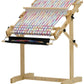 The Schacht Flip Folding Rigid Heddle Loom from Schacht Spindle Co. stands on a sturdy wooden base. The loom displays multicolored fabric featuring diagonal stripes in blue, red, yellow, and white. Several adjustable knobs and levers are visible for modifying settings. The plain white background provides clarity for warping and weaving instructions.