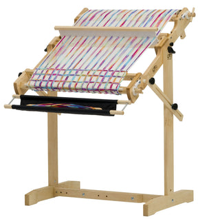 The Schacht Flip Folding Rigid Heddle Loom from Schacht Spindle Co. stands on a sturdy wooden base. The loom displays multicolored fabric featuring diagonal stripes in blue, red, yellow, and white. Several adjustable knobs and levers are visible for modifying settings. The plain white background provides clarity for warping and weaving instructions.