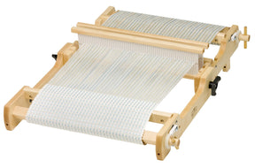 The Schacht Flip Folding Rigid Heddle Loom from Schacht Spindle Co. showcases white and light blue threads partially woven into fabric on a wooden tabletop. This loom includes a heddle, tension knobs, and side supports, and is set against a white background. It’s perfect for those who are following warping and weaving instructions for intricate patterns.