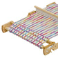 A Schacht Flip Folding Rigid Heddle Loom by Schacht Spindle Co., adorned with colorful threads in stripes of various hues, is actively being used to create a partially completed woven fabric. The plain white background serves to highlight the vibrant threads and detailed wooden construction of the two-heddle weaving setup.