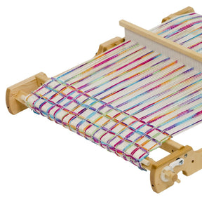 A Schacht Flip Folding Rigid Heddle Loom by Schacht Spindle Co., adorned with colorful threads in stripes of various hues, is actively being used to create a partially completed woven fabric. The plain white background serves to highlight the vibrant threads and detailed wooden construction of the two-heddle weaving setup.