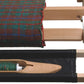 A close-up of a wooden loom with a fabric in progress, showcasing a multicolored plaid pattern in shades of green, brown, red, and blue. Below the fabric, resting in the Schacht Flip Trap by Schacht Spindle Co., is a wooden shuttle with green thread placed in a black fabric compartment.