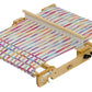 The Schacht Flip Folding Rigid Heddle Loom by Schacht Spindle Co. boasts multicolored threads stretched across its frame, which is equipped with pegs and adjustable mechanisms for precise tensioning of the threads. At its center is a comb-like reed to assist in the weaving process, making it an ideal tool for both beginners and those following warping and weaving instructions.