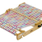 A Schacht Spindle Co. Schacht Flip Folding Rigid Heddle Loom with a partially completed colorful fabric, featuring intricate multicolored stripes. The loom’s frame is natural wood, and several mechanisms are visible, including rollers and tension adjustments, holding the warp threads tight.