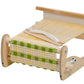 The Schacht Cricket Rigid Heddle Loom by Schacht Spindle Co. is a small, portable weaving loom with a natural wood finish that showcases a piece of fabric with a checkered green and white pattern in progress. The loom's threads are taut, and a shuttle with white yarn is placed on top, making it perfect for intricate weaving projects.