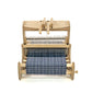 A Schacht Cricket 15" Quartet from Schacht Spindle Co. features a partially woven fabric in a blue and white checkered pattern. The setup is minimal, highlighting the loom's various components such as warp threads, heddles, and reed as they work together on this weaving project.