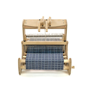 A Schacht Cricket 15" Quartet from Schacht Spindle Co. features a partially woven fabric in a blue and white checkered pattern. The setup is minimal, highlighting the loom's various components such as warp threads, heddles, and reed as they work together on this weaving project.