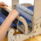 A person operates a Schacht Cricket 15" Quartet loom from Schacht Spindle Co., weaving with blue and white threads. Their hands skillfully adjust the thread and use the shuttle to create a patterned fabric. The loom's components and the partially woven fabric are clearly visible, beautifully showcasing its 15-inch weaving width.