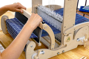 A person operates a Schacht Cricket 15" Quartet loom from Schacht Spindle Co., weaving with blue and white threads. Their hands skillfully adjust the thread and use the shuttle to create a patterned fabric. The loom's components and the partially woven fabric are clearly visible, beautifully showcasing its 15-inch weaving width.