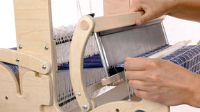 A person weaving fabric on a Schacht Cricket 15" Quartet by Schacht Spindle Co. Their hands are adjusting the threads and shuttle, creating a blue and white patterned fabric. The loom's structure and threads are clearly visible in the image, with a weaving width accommodating intricate designs.