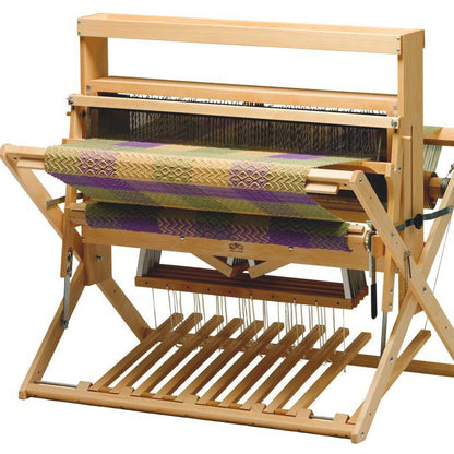 A versatile Schacht 36" Mighty Wolf Floor Loom from Schacht Spindle Co. with a multi-colored woven fabric partially completed. The fabric features an intricate pattern with green, purple, and beige colors. This 36" jack loom is equipped with various levers, strings, and beams essential for the weaving process.