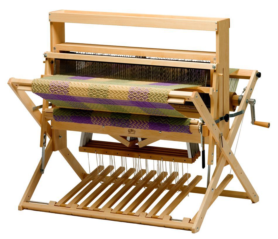 A Schacht 36" Mighty Wolf Floor Loom by Schacht Spindle Co. with an intricate tapestry in green, yellow, and purple being woven. The versatile weaving loom is equipped with levers, beams, and pedals for creating various patterns and textures in fabric.